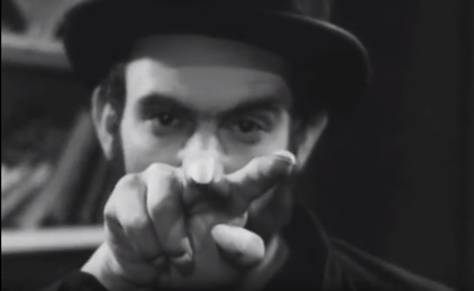 Black & white movie still of a man wearing a top hat and a cowl, glaring at the camera and pointing two creepy long fingers.